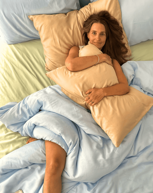Woman in bed holding peach cotton top sheet set with blue duvet, light green sheet visible underneath