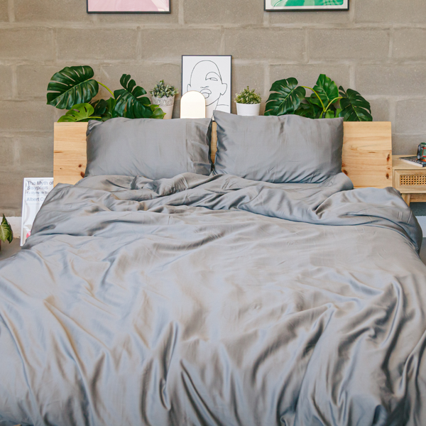 Cama de casal com roupa de cama cinzenta clara em tecido liso, ligeiramente amarrotada, com duas almofadas a condizer. A cabeceira é de madeira clara com várias plantas verdes e pequenos vasos decorativos apoiados por cima, sobre um fundo de parede em betão.