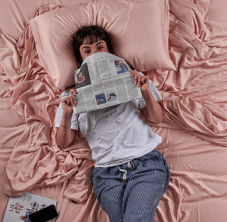 Woman lying on pink bamboo bed sheet reading newspaper, overhead view