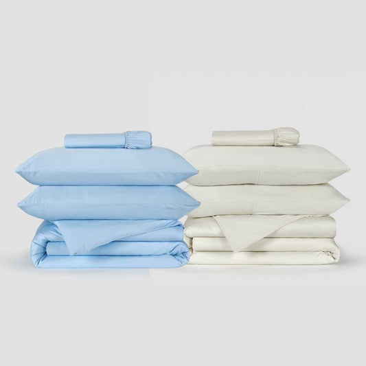 Two Momo Cotton duvet cover sets displayed side by side, one light blue and one cream, with folded sheets and rolled linens