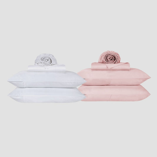 White and pink bed sheet sets stacked with pillows and rolled duvet on neutral background