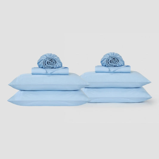 Light blue cotton sheet set displayed with pillows and folded linens on white background