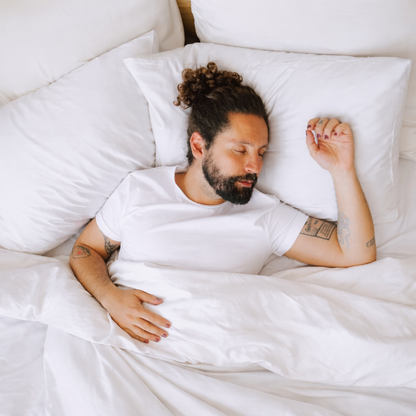 Homem a dormir tranquilamente numa cama com lençóis brancos, envolvido em roupa de cama minimalista e confortável, transmitindo sensação de descanso profundo e bem-estar.