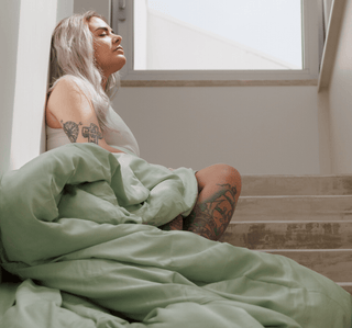 Woman wearing cream bamboo duvet cover set in sage green, seated by window showing soft fabric texture and drape.